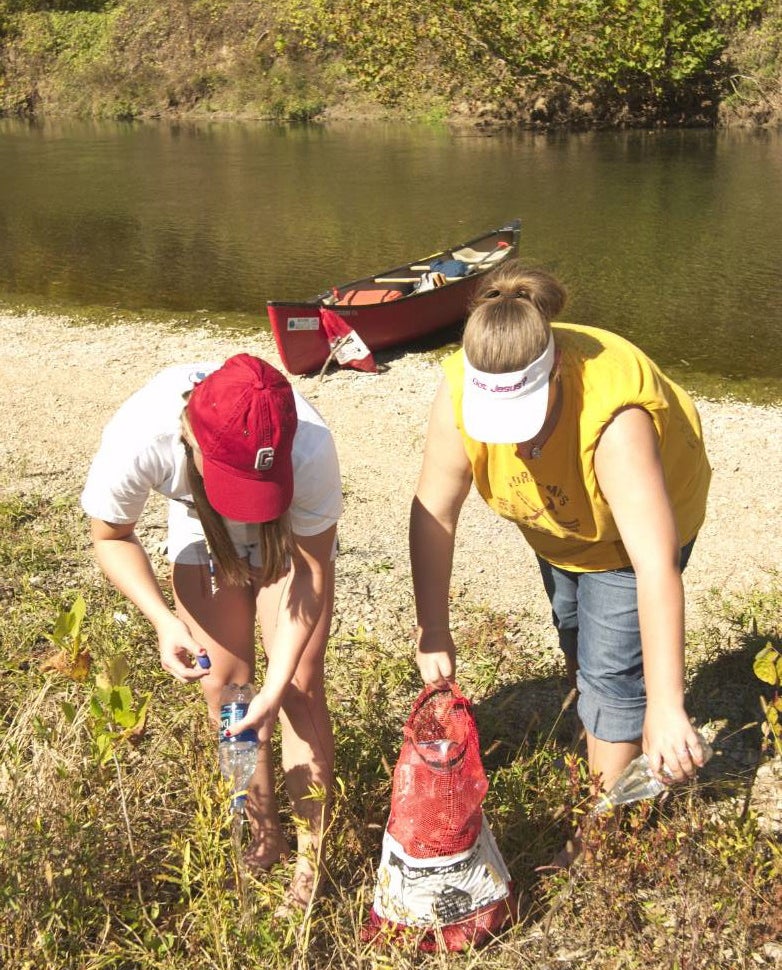 Two canoers put plastic containers in a "Stash Your Trash" red mesh bag.