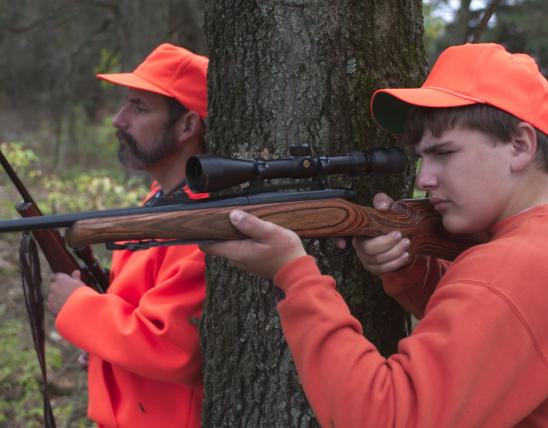 Two hunters in safety orange sit by a tree. One is taking aim at a deer.